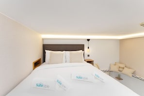 Habitación de hotel con una cama bien hecha, una mesita de noche, una lámpara y una pequeña zona de estar con cojines.