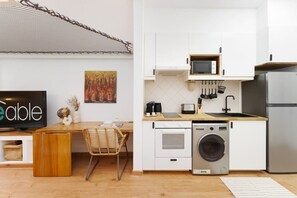 A compact kitchen with white cabinets, a microwave, a washing machine, and a dining area with a wooden table and chairs. - Extraordinary Loft Living in JVC by Suiteable (Dubai)