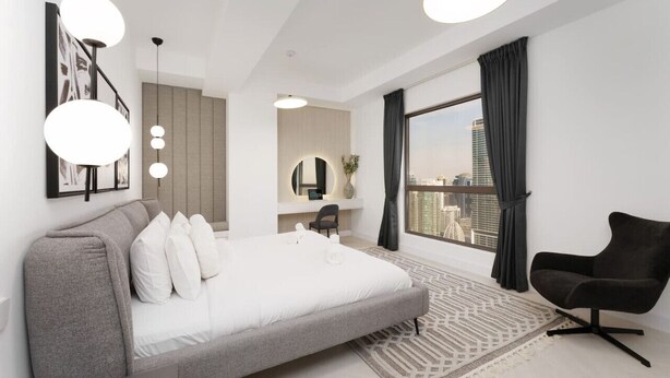 Ein modernes Schlafzimmer mit einem großen Bett, einem Schreibtisch, einem Sessel und Blick auf die Stadt durch ein großes Fenster.