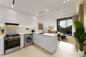 A modern kitchen with white cabinetry, a built-in stove, and a washing machine. There is a large window providing a view of the city. - Premium Living Apartment in JVC by Suiteable (Dubai)