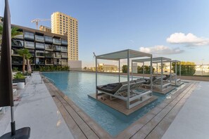 A rooftop pool with lounge chairs and a covered seating area, surrounded by modern buildings and palm trees.
