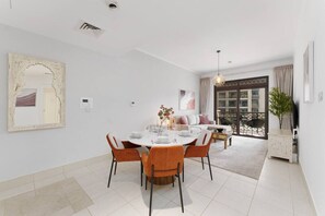 Ein Essbereich mit einem runden Tisch, gedeckt für vier Personen, umgeben von orangefarbenen Stühlen. An der Wand befindet sich ein großer Spiegel, ein Deckenlüfter und eine Tür, die auf einen Balkon mit Gartenmöbeln führt.