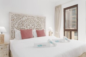 Ein Hotelzimmer mit einem großen Bett, weißer Bettwäsche, einem dekorativen Kopfteil, zwei Nachttischlampen und einem Fenster mit Vorhängen.