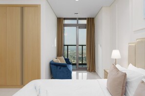 Ein Hotelzimmer mit einem großen Bett, einem blauen Sessel, einem Nachttisch mit Lampe und Blick auf die Stadt durch ein großes Fenster.
