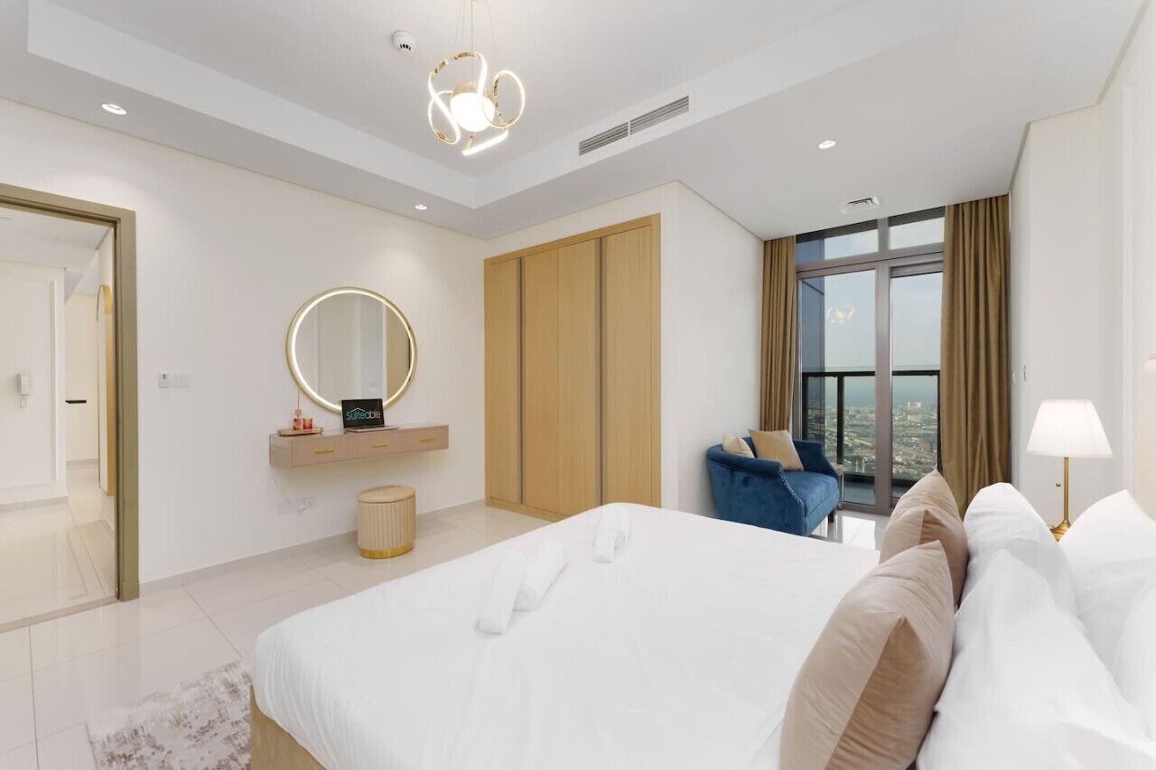Una habitación de hotel moderna con una cama grande, un espejo redondo, un armario, una zona de estar con un sofá azul y vistas a la ciudad a través de amplias ventanas.