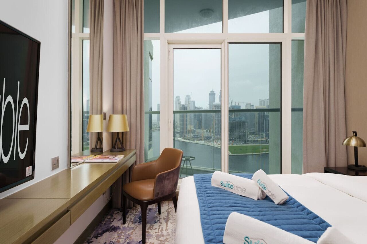 A hotel room with a large window offering a city view, a bed with a blue quilt, a desk with a lamp, and a chair.