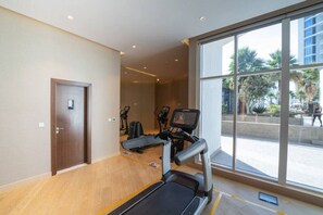 Un gimnasio con caminadora, bicicletas estáticas y un amplio ventanal que ofrece vistas a palmeras y un edificio.