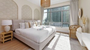 Une chambre d’hôtel moderne avec un grand lit, une table de chevet, un fauteuil en osier, un canapé blanc et une grande fenêtre donnant sur la ville.