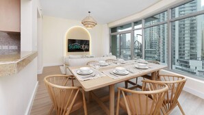 Un espace de salle à manger moderne avec une grande table dressée pour six personnes, une cuisine dotée d’un îlot central et une grande fenêtre offrant une vue sur la ville.