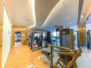Academia moderna com diversos equipamentos de exercício, piso de madeira e amplas janelas que permitem entrada de luz natural.