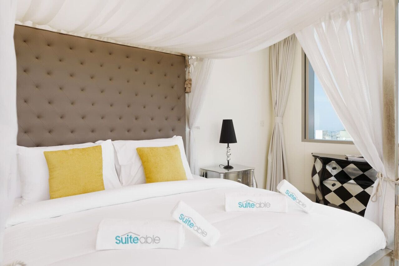 Una habitación de hotel con un cabecero capitonado, una cama con ropa blanca y almohadas amarillas, una mesita de noche con una lámpara y una ventana con cortinas translúcidas.