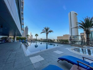 A rooftop pool with clear blue water, surrounded by lounge chairs and palm trees.