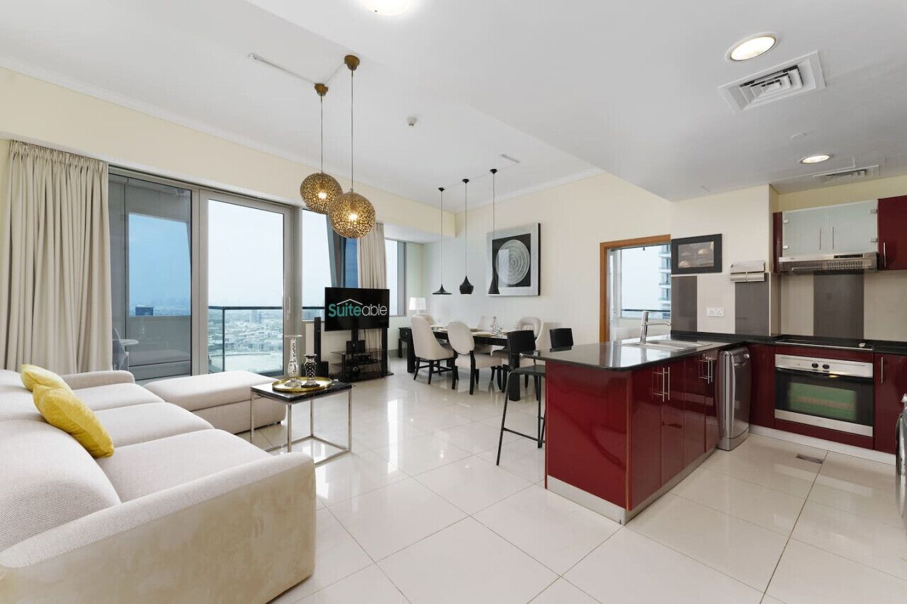 A modern living room with a sofa, a dining area with chairs, a kitchen with red cabinets, and a large window with curtains.