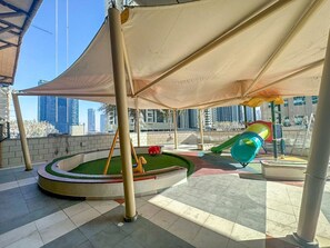 Um parquinho com escorrega e balanços, cercado por uma área arborizada com vista para edifícios ao fundo.