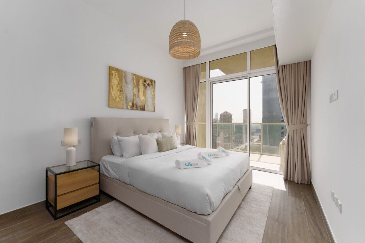 A modern bedroom with a large bed, a bedside table, a hanging light fixture, and a view of the city through large windows.