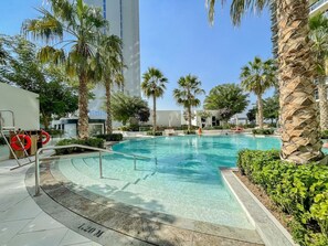 Ein Swimmingpool mit klarem Wasser, umgeben von Palmen und einem modernen Gebäude im Hintergrund.