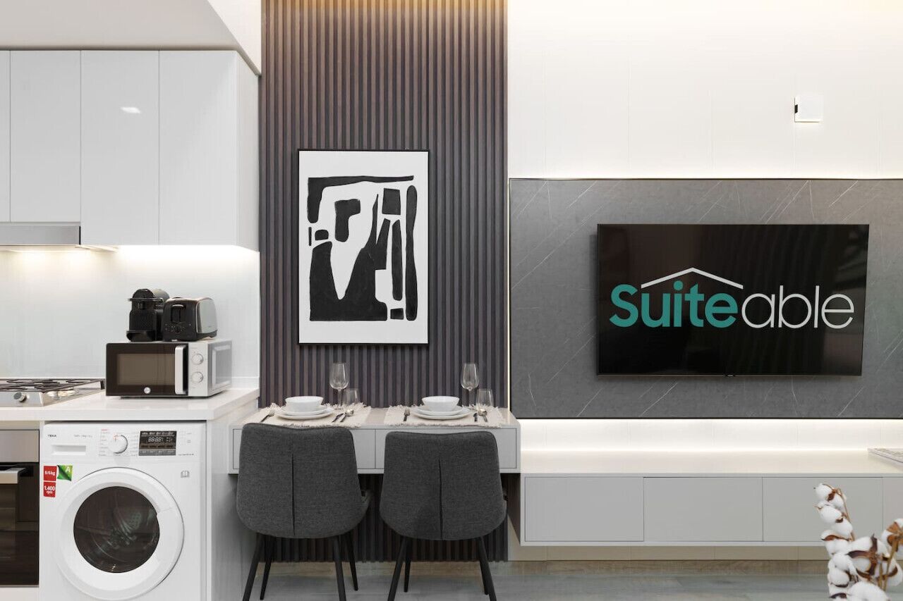Ett modernt kök med mikrovågsugn, brödrost och tvättmaskin, en matplats med ett bord för två personer och en stor platt-TV som är monterad på väggen och visar ordet "SuiteAble".