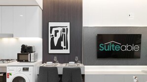 A modern kitchen with a microwave, toaster, and washing machine, a dining area with a table set for two, and a large flat-screen TV mounted on the wall displaying the word "SuiteAble". - Luxury Redefined Studio in AHAD Residence (Business Bay)