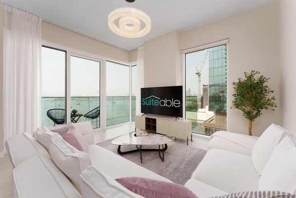 Un salon moderne doté d’un grand téléviseur à écran plat, de canapés blancs, d’une table basse et de grandes fenêtres offrant une vue sur la mer.