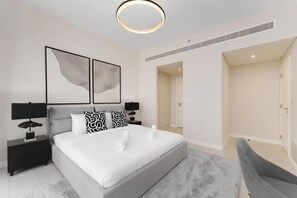 Une chambre à coucher moderne avec un grand lit, deux tables de chevet avec des lampes, un fauteuil à revêtement gris et des œuvres d’art abstraites accrochées au mur.