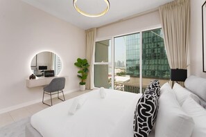 A modern hotel room with a large bed, a round mirror, a desk with a computer, a potted plant, and a view of a cityscape through large windows.