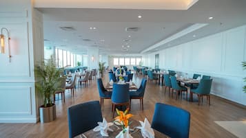 Un comedor amplio con varias mesas y sillas, ventanas grandes y plantas en macetas.