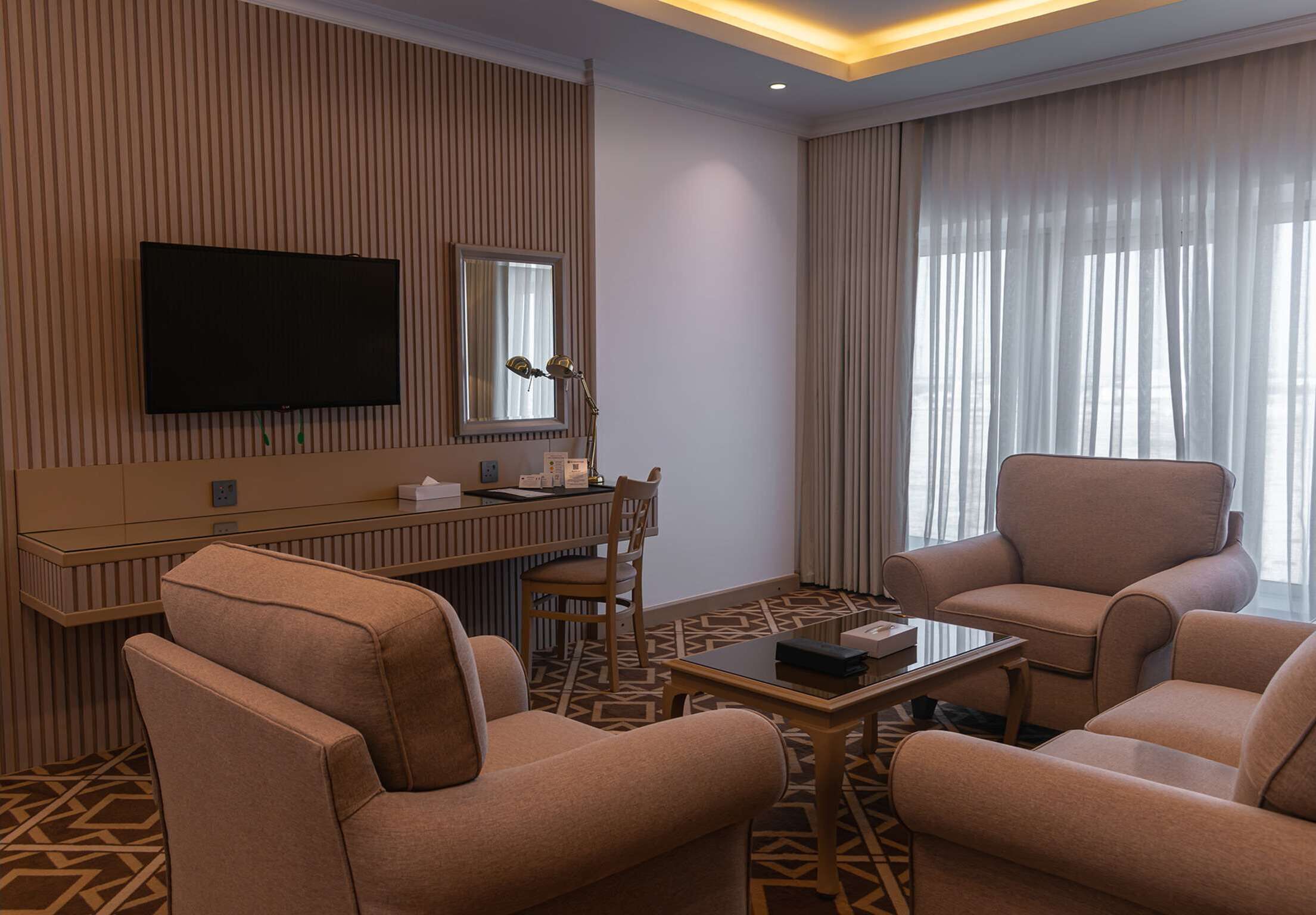 Hotelkamer met een wandgemonteerde flatscreen televisie, een bureau met stoel, een bank, een salontafel en een groot raam met gordijnen.