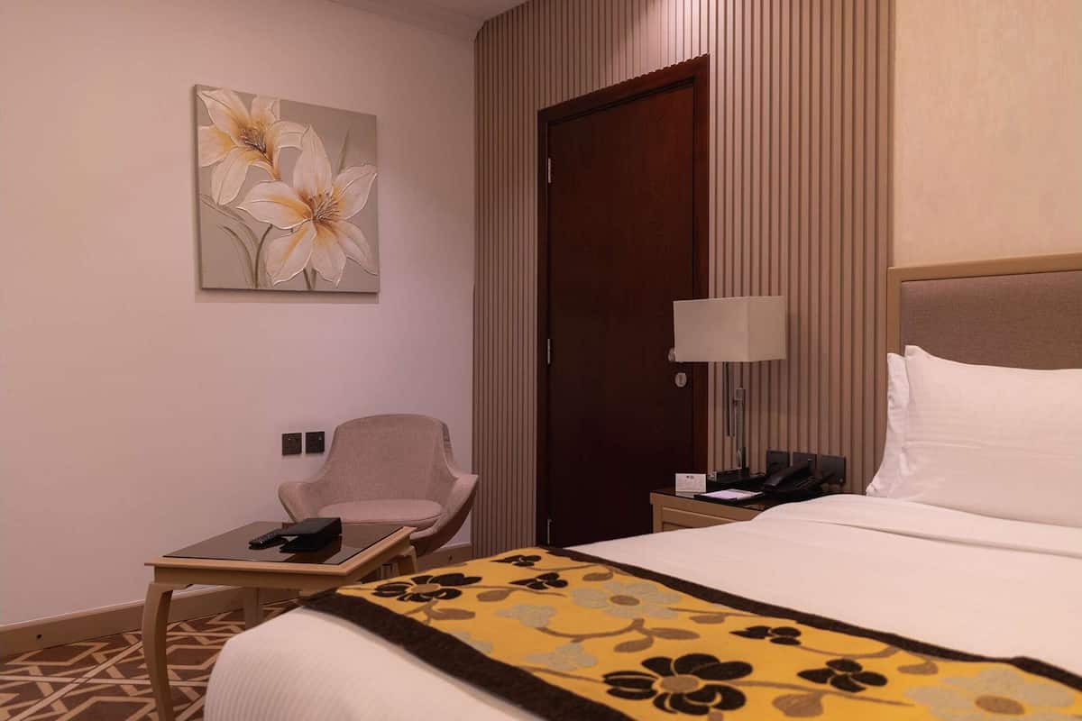 A hotel room with a bed, a chair, a small table, a lamp, and a painting on the wall.