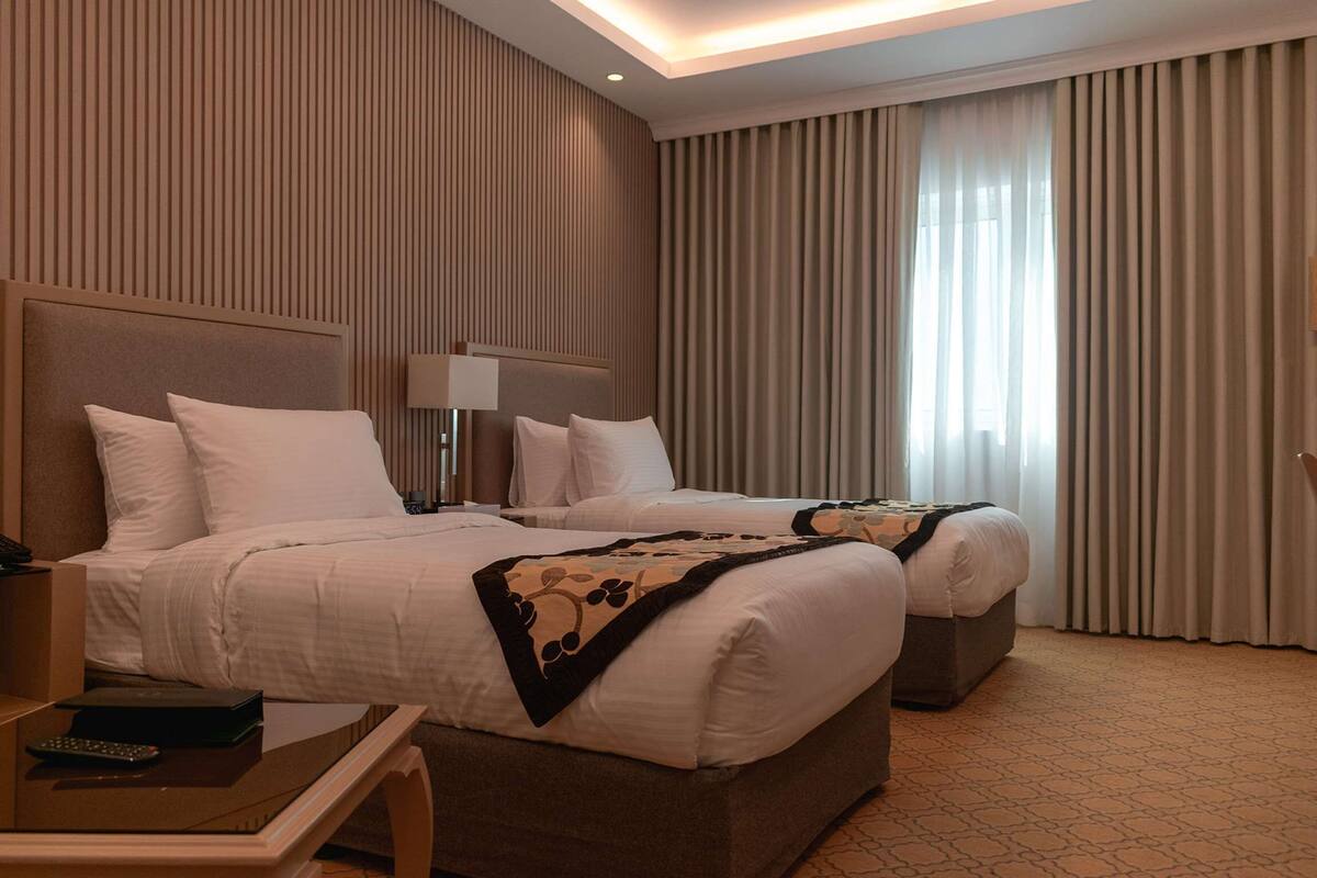 A hotel room with two beds, a bedside table with a lamp, and a large window with curtains.