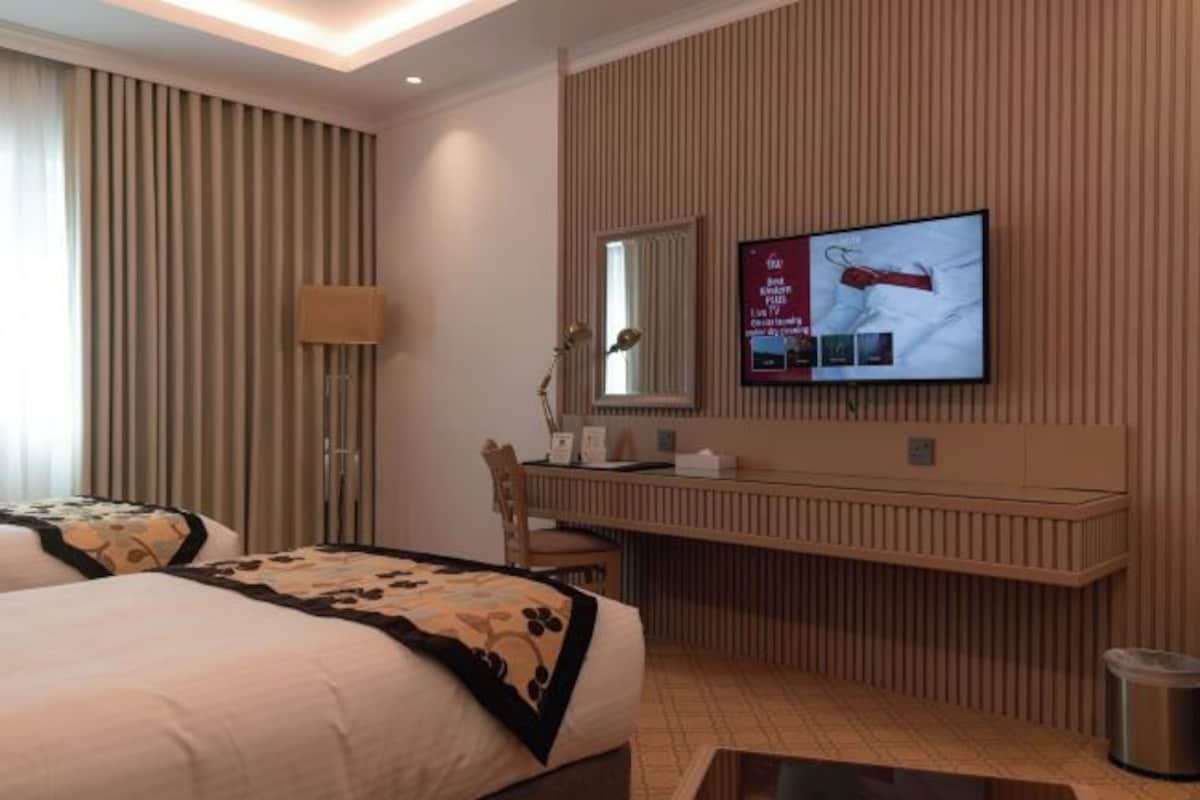 A hotel room with two beds, a wooden panel wall, a TV mounted above a wooden desk, and a small trash bin.