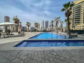 A swimming pool with lounge chairs and umbrellas, surrounded by palm trees and high-rise buildings.