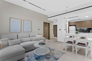 A modern living room with a grey sofa, a coffee table, and a dining area with white chairs and a table. There are framed artworks on the wall and a kitchen area in the background.