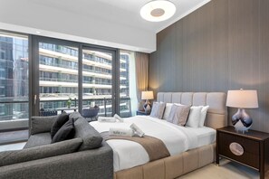 A modern hotel room with a large bed, a sofa, a nightstand, and a view of a high-rise building.