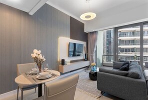 A modern living room with a dining area, a grey sofa, a flat-screen TV, and a large window with a city view.