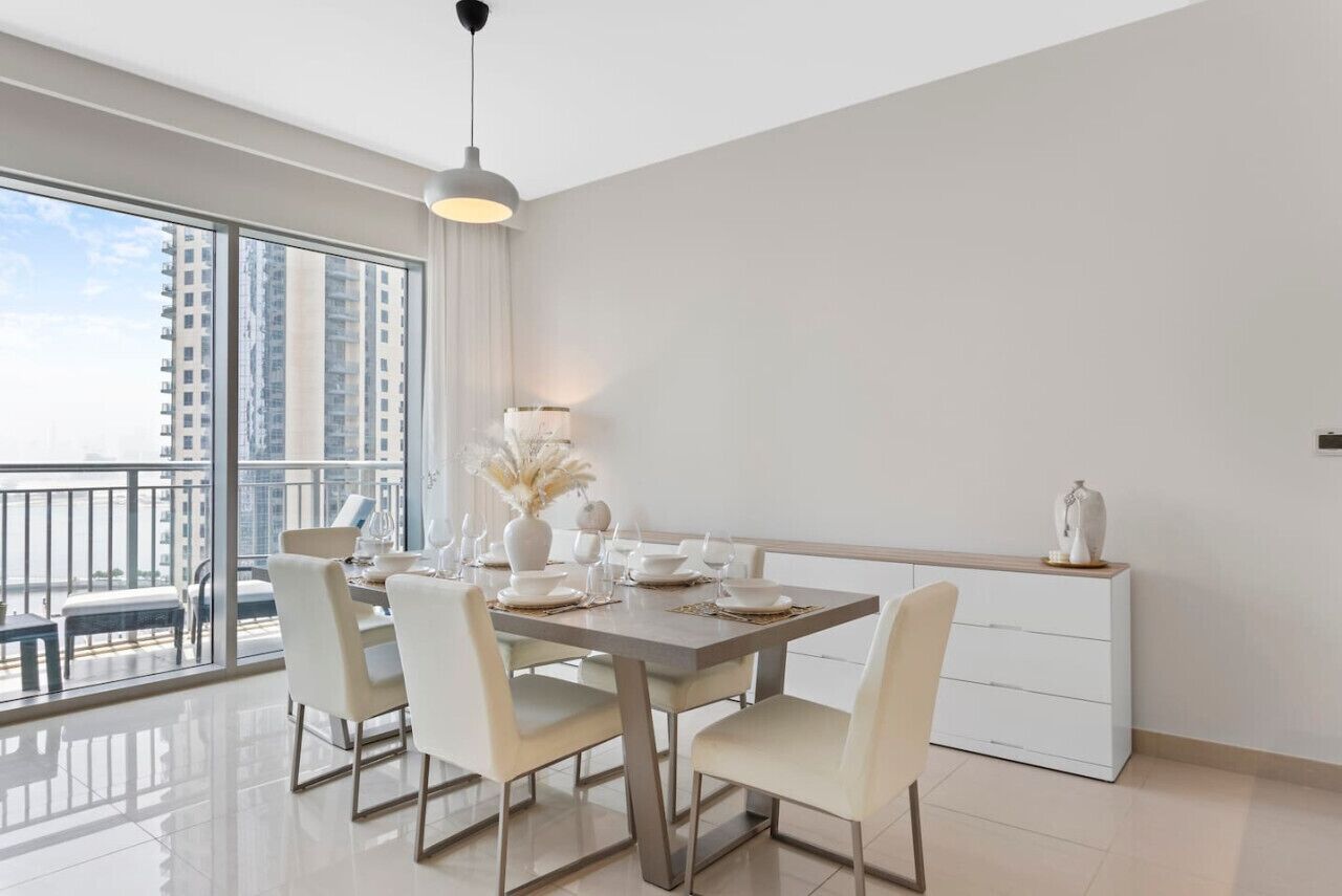 A modern dining area with a large window, a dining table set with white plates and glasses, and a white cabinet against the wall.