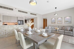 A modern dining area with a long table set for four, a kitchen in the background, and a living area with a sofa and artwork on the wall.