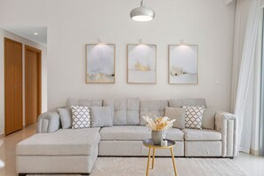 A modern living room with a grey sectional sofa, a small round table with a vase, and three framed abstract paintings on the wall. - Luxurious Living 3 Bedroom at Harbour Views (Dubai)