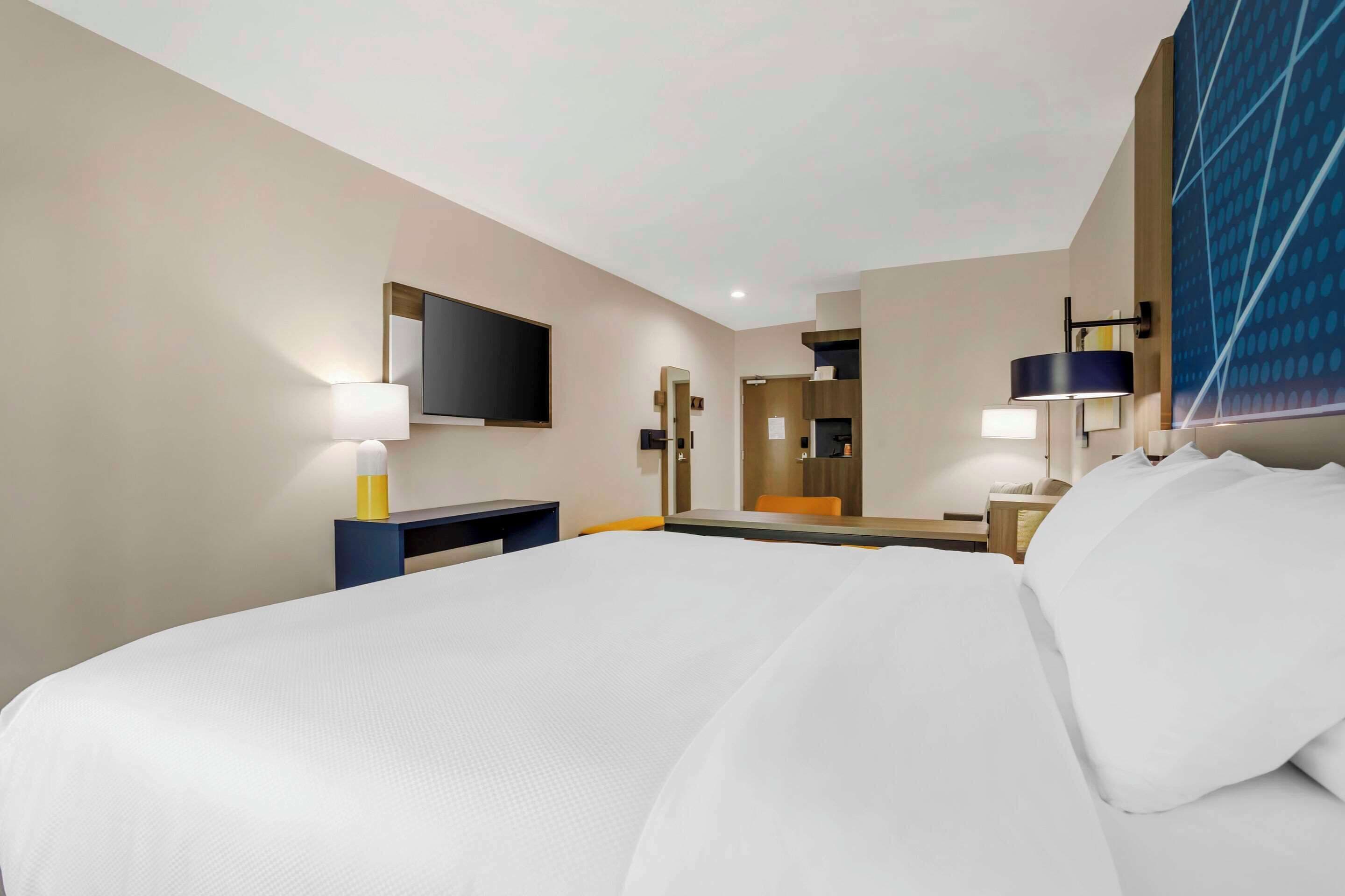 Habitación de hotel con una cama grande, un escritorio, una silla, un televisor de pantalla plana, un espejo y una lámpara fijada a la pared.