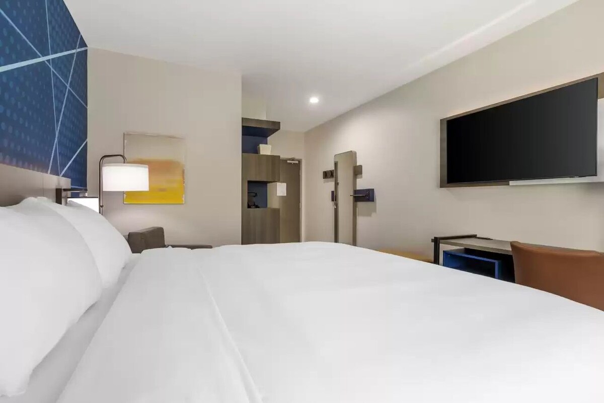 A hotel room with a large bed, a desk, a chair, a TV, and a wall-mounted mirror.