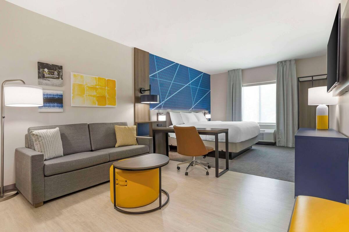 A hotel room with a sofa, a small round table, a desk with a chair, a bed, and a TV.