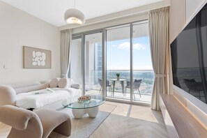 A modern hotel room with a sofa, a glass coffee table, a flat-screen TV, and a balcony with a view of the city.