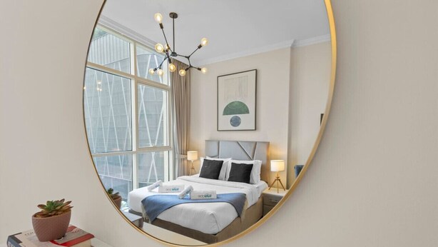 Ein modernes Hotelzimmer mit großem Spiegel, der das Bett, Nachttischlampen, ein Fenster mit Vorhängen und eine Topfpflanze reflektiert.