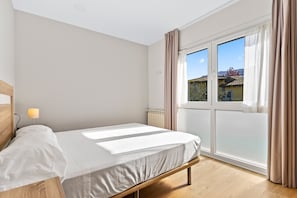 Une chambre à coucher comprenant un lit, une table de chevet, une lampe, une fenêtre avec des rideaux et une vue sur les bâtiments à l’extérieur.