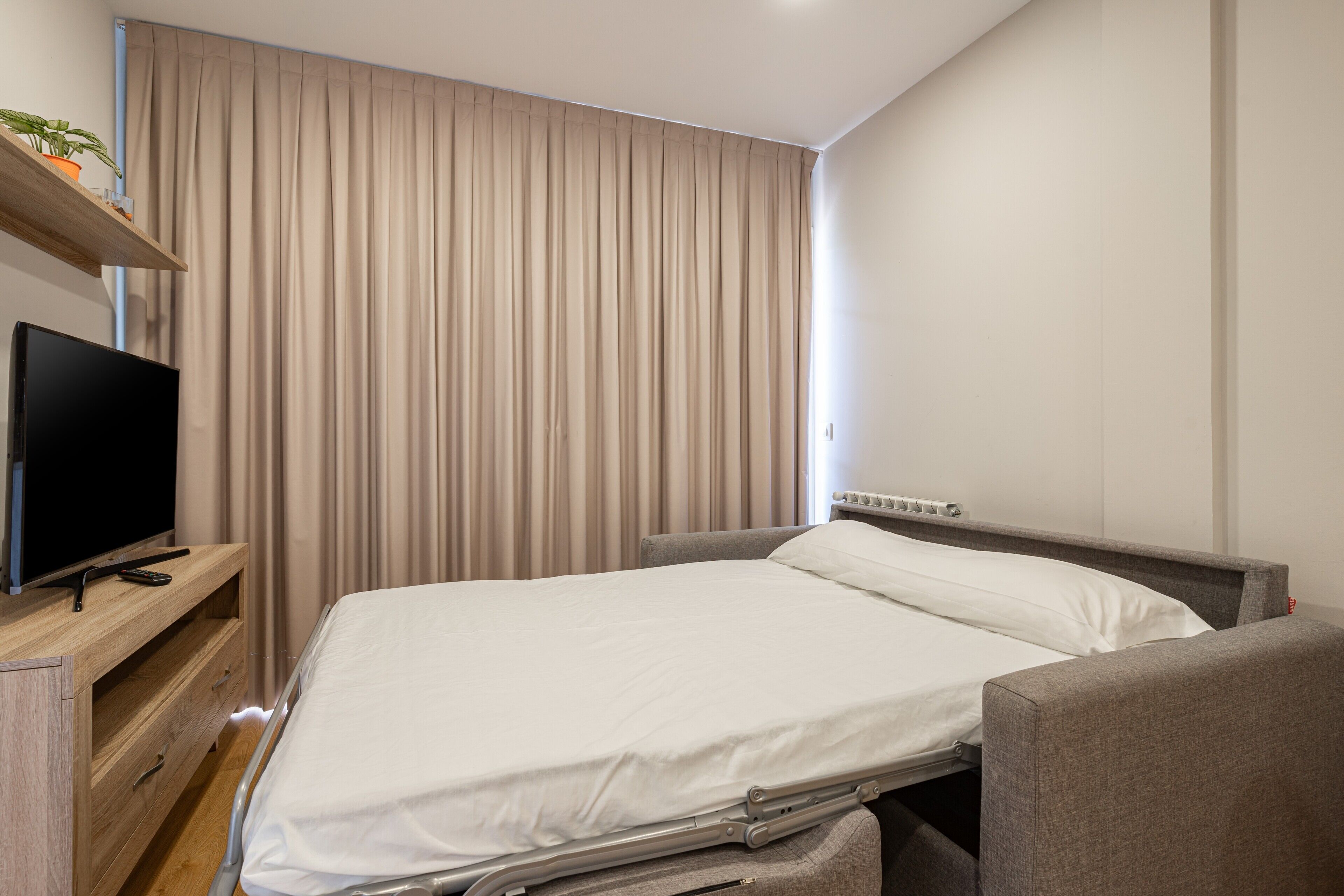 A hotel room with a bed, a TV on a wooden dresser, and a curtain.
