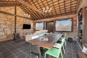 A rustic room with a stone wall, wooden beams, a fireplace, a sofa, a dining table with chairs, and a television.