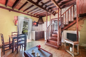 A rustic interior with a wooden staircase, a dining area with chairs and a table, a kitchen with a stove, and a television on a stand.