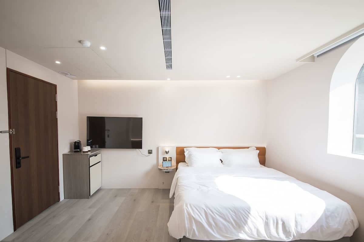 Un dormitorio moderno con puerta de madera, televisión de pantalla plana, mesita de noche con lámpara y una cama grande.