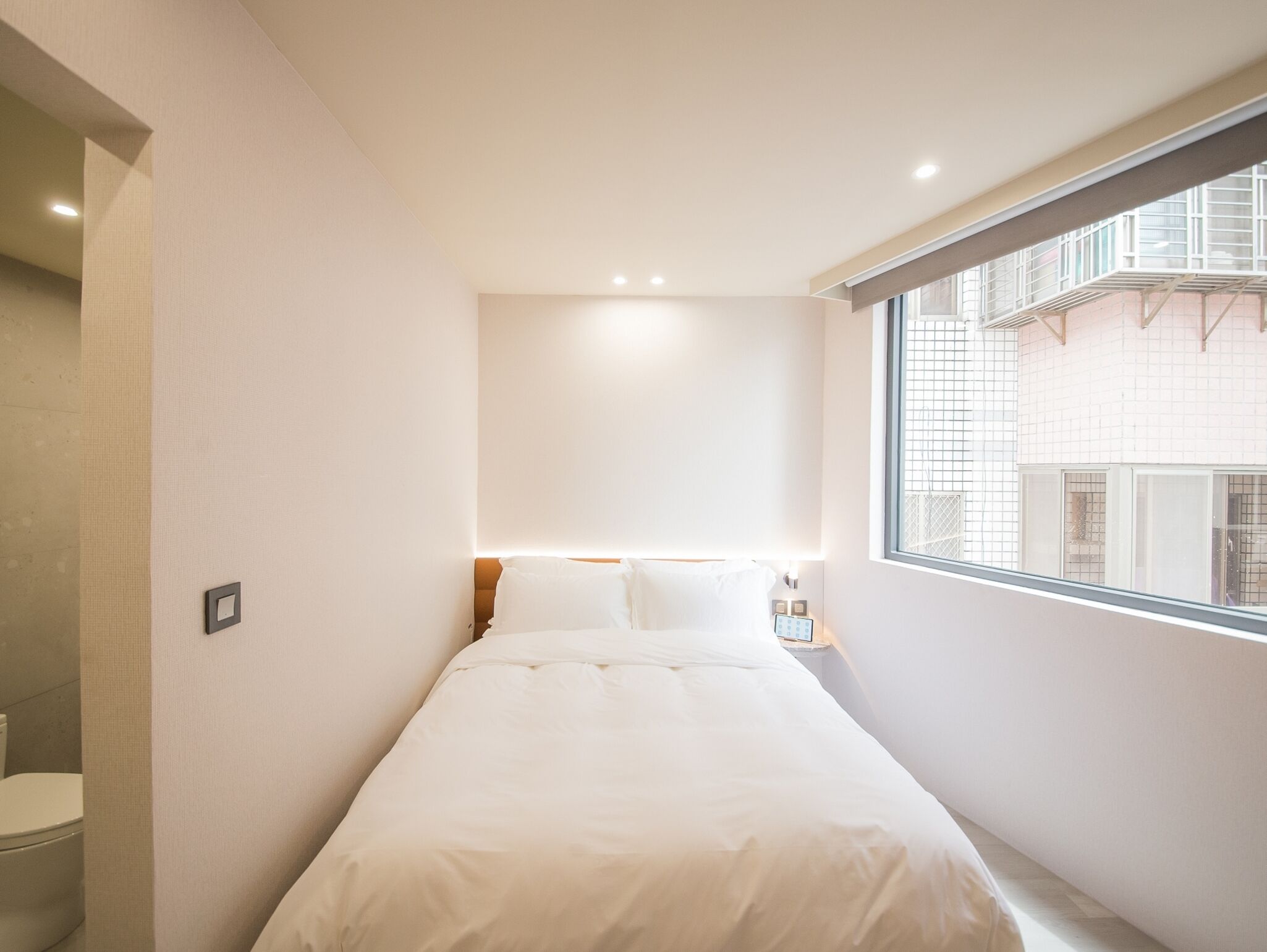 Een moderne hotelkamer met een bed, een klein nachtkastje, een raam met uitzicht op gebouwen en een badkamer met toilet.