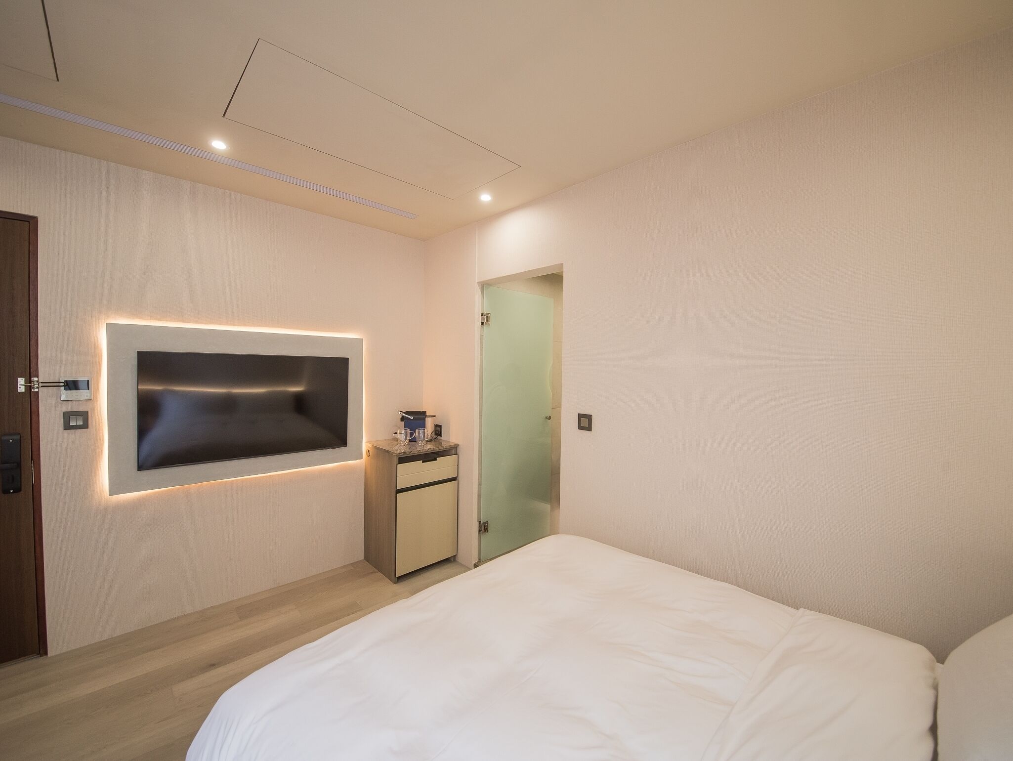 Una habitación de hotel moderna con una cama grande, un televisor de pantalla plana montado en la pared, una mesita de noche pequeña y una puerta que da a otra habitación.