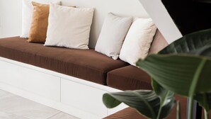 Een bruine bank met witte en beige kussens, een wit kastje en een grote groene plant.